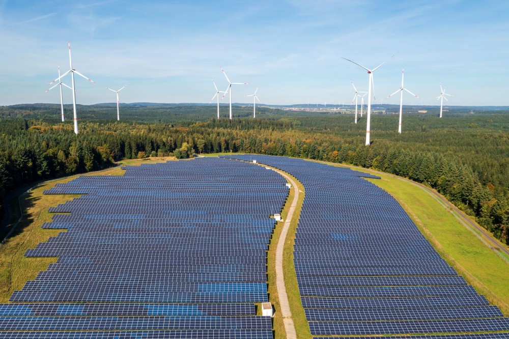 Solar panels in a field surrounded by trees under a vast blue sky with wind turbines in the distance