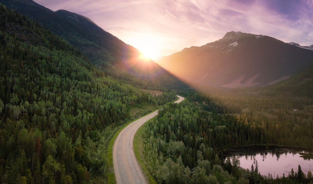 Photo of the sun setting behind lush green mountains with a winding road