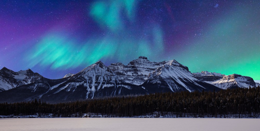 Photo of the aurora borealis lighting up the sky beneath a snow covered mountain range in Canada