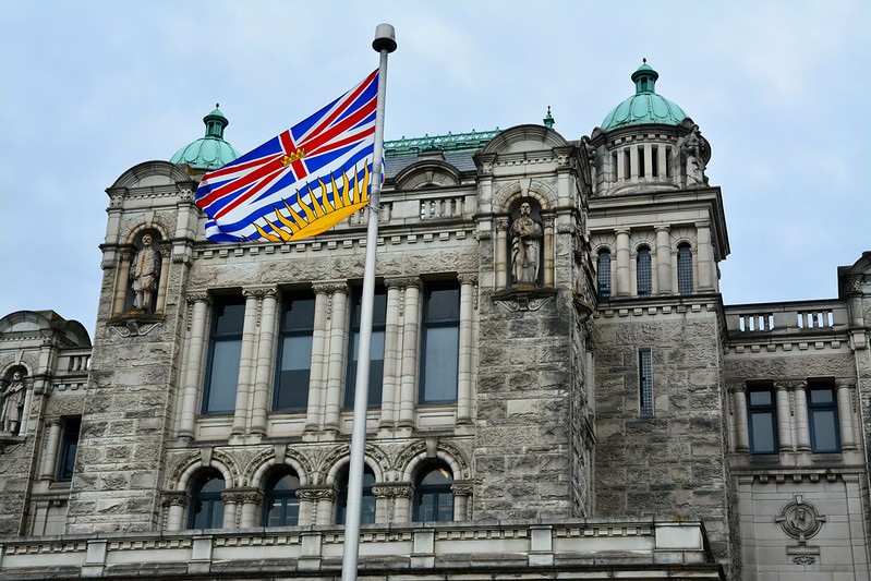 The front facade of the legislature building of B.C. with the provincial flag flying in the foreground