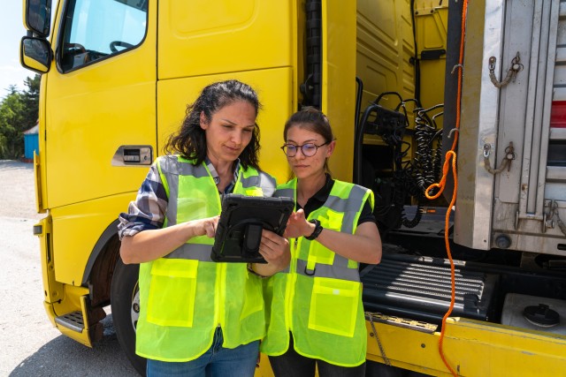 Two women in yellow safety vests standing in front of a large yellow truck checking information on a tablet