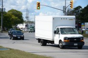 A white box truck drives down a road lined with businesses in the background