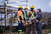 Four construction workers huddle together as they take a quick break and plan out their next set of tasks. They are all dressed in workwear and proper safety gear.